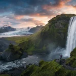 Paesaggio epico dell'Islanda al tramonto: un'imponente cascata lungo una scogliera verdeggiante, una spiaggia di sabbia nera e un ghiacciaio sullo sfondo