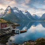 Paesaggio di viaggio in Norvegia con fiordi e montagne innevate che si riflettono nelle acque azzurre, una tipica cabina rossa (rorbue) sulla riva rocciosa e una barca.