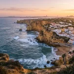 Fotografia panoramica di viaggio in Portogallo: un paesaggio costiero con falesie dorate dell'Algarve, le onde dell'Oceano Atlantico, un tradizionale villaggio portoghese con case bianche e dettagli colorati.