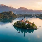 Rappresentazione aerea del Lago di Bled in Slovenia all'alba con Chiesa dell'Assunzione, il Castello di Bled arroccato sulla collina in lontananza, sullo sfondo delle Alpi Giulie.