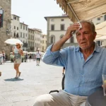 Uomo seduto in un caffè all'aperto si asciuga il sudore dalla fronte con un fazzoletto durante una giornata di caldo record con un termometro digitale sullo sfondo che mostra 42 gradi.