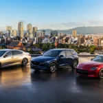 Auto giapponesi disposti su piazzale panoramico in Giappone, sullo sfondo una città contemporanea con elementi architettonici e montagne
