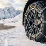 Primo piano del pneumatico di un'auto montato con catene da neve su una strada di montagna innevata, con montagne coperte di neve sullo sfondo.