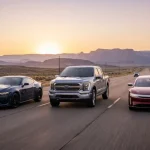 Auto americane, su una grande highway degli Stati Uniti al tramonto: moderna muscle car sportiva affiancata da un grande pick-up e da una moderna automobile elettrica dal design tecnologico.
