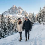 Una coppia cammina su un sentiero di montagna in inverno tra boschi innevati con sullo sfondo una cima dolomitica.