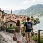 Una coppia rilassata durante delle vacanze brevi cammina su una strada di un borgo italiano affacciato su un lago, sullo sfondo montagne verdi e un centro storico con edifici in pietra.