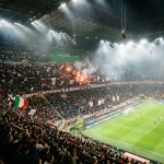 Una fotografia panoramica dall'alto, ricca di atmosfera, mostra un moderno stadio gremito di tifosi appassionati durante una partita serale dei campionati di calcio italiani.