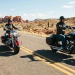 Due motociclisti viaggiano su moto americane custom, una nera e una rossa, lungo una strada deserta tra rocce rosse e cactus.