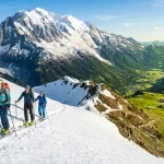 Immagine di fine stagione sciistica in quota con sciatori su una pista innevata, circondati da cime ancora bianche e maestose e prati verdi a valle