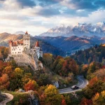 Veduta autunnale panoramica del Castello di Bran in Romania, arroccato su una rupe e circondato da fitte foreste di alberi con foglie rosse e arancioni.