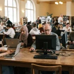 Fotografia sul tema del digital divide con persone diverse per età e condizioni sociali all'interno di una biblioteca pubblica che utilizzano dispositivi.